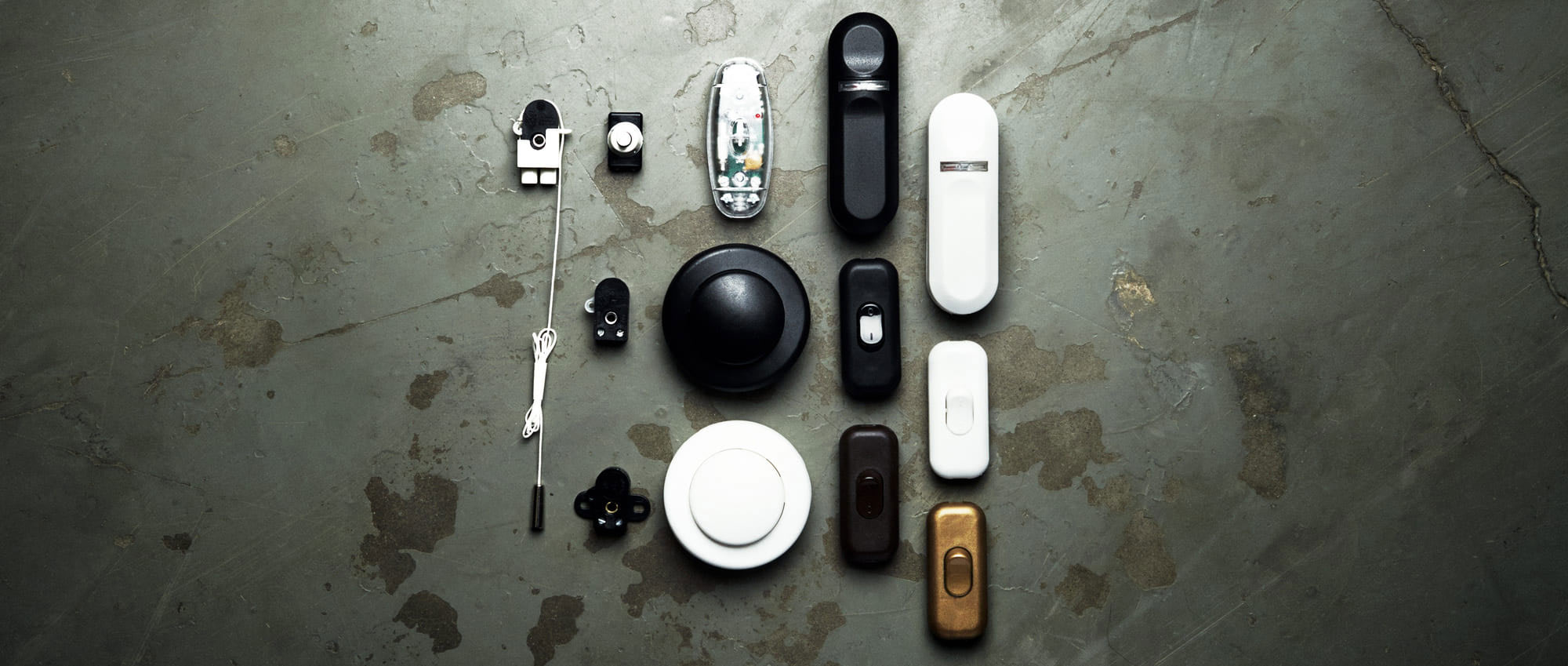 Översiktsbild på ett sortiment av strömbrytare, dimmers och stickproppar i olika former, färger och material – samlade på ett betonggolv i dekorativ layout.