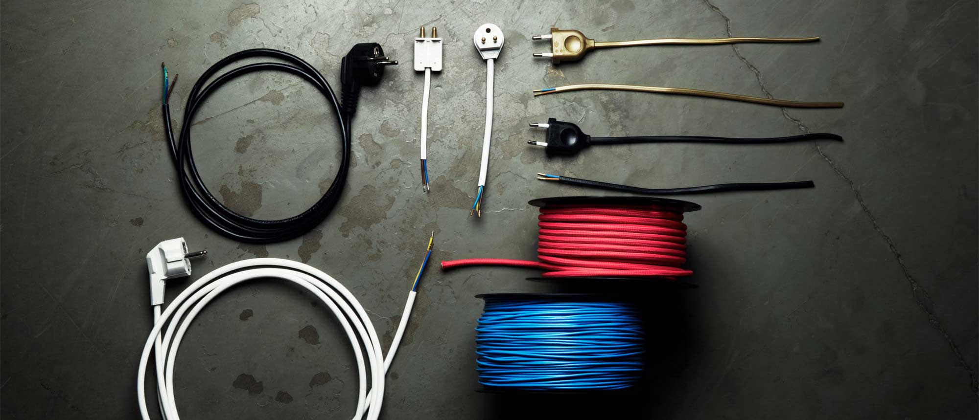 Olika typer av sladdställ och textilkablar med stickproppar och rullar i svart, vitt, guld, rött och blått, liggande på ett betonggolv.