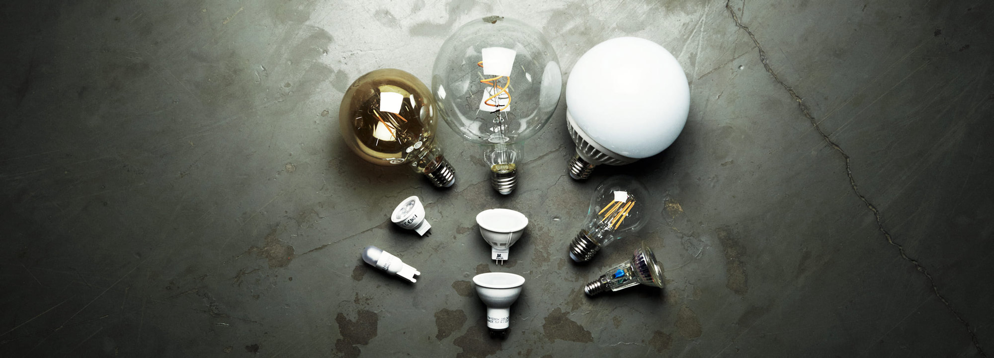 Olika typer av LED-ljuskällor i varierande former och socklar uppställda på ett betonggolv.