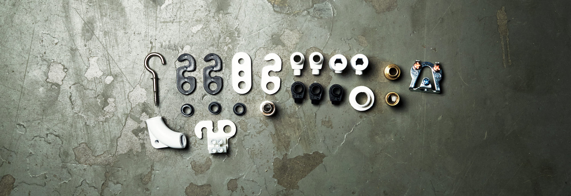Olika monteringstillbehör för belysning, innehåller krokar, dragavlastningar, genomföringsdetaljer och kopplingsplintar, upplagda i rad på en betongyta.
